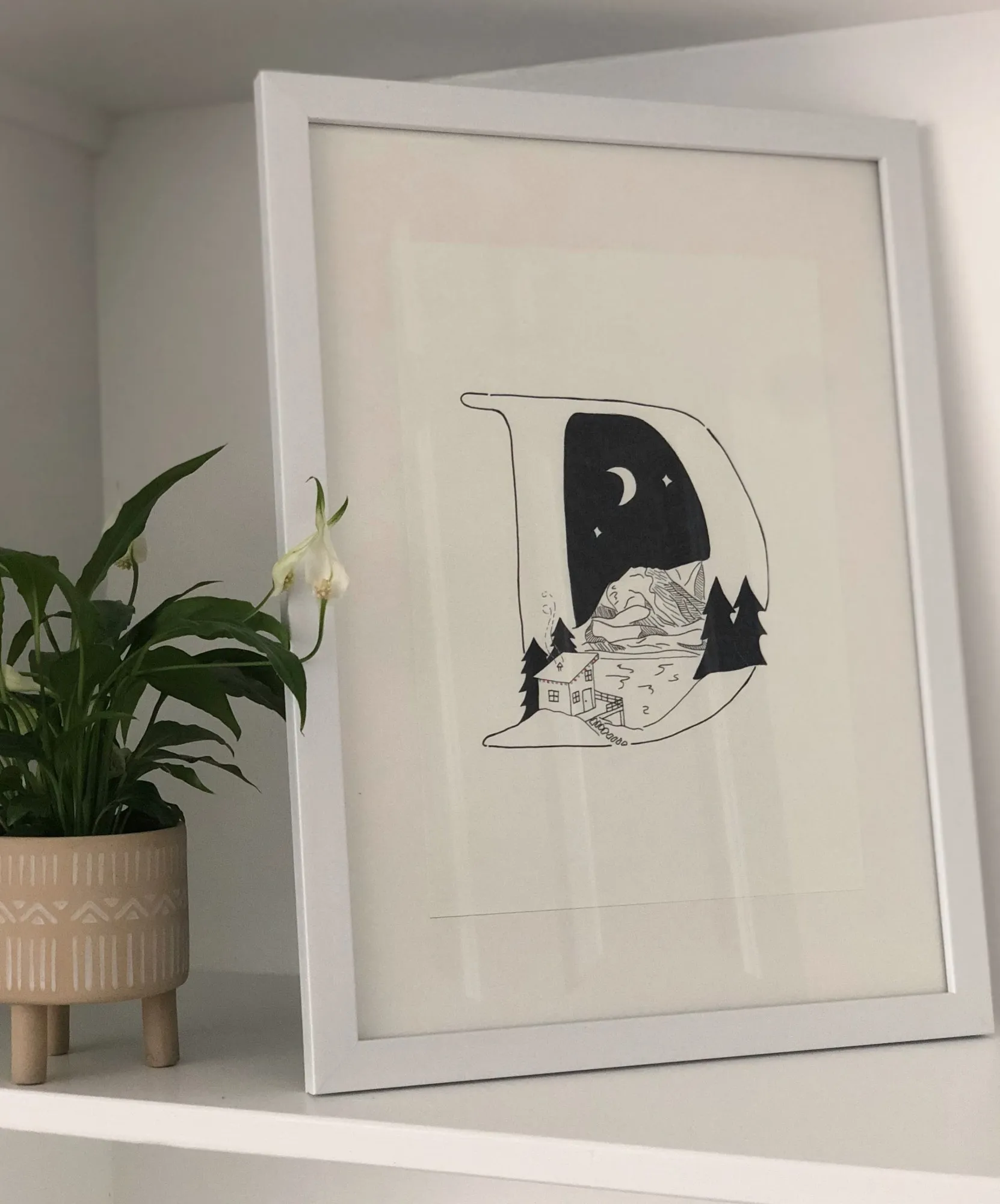 A framed fine-liner drawing of the letter D with a cabin, pine trees and a crescent moon inside