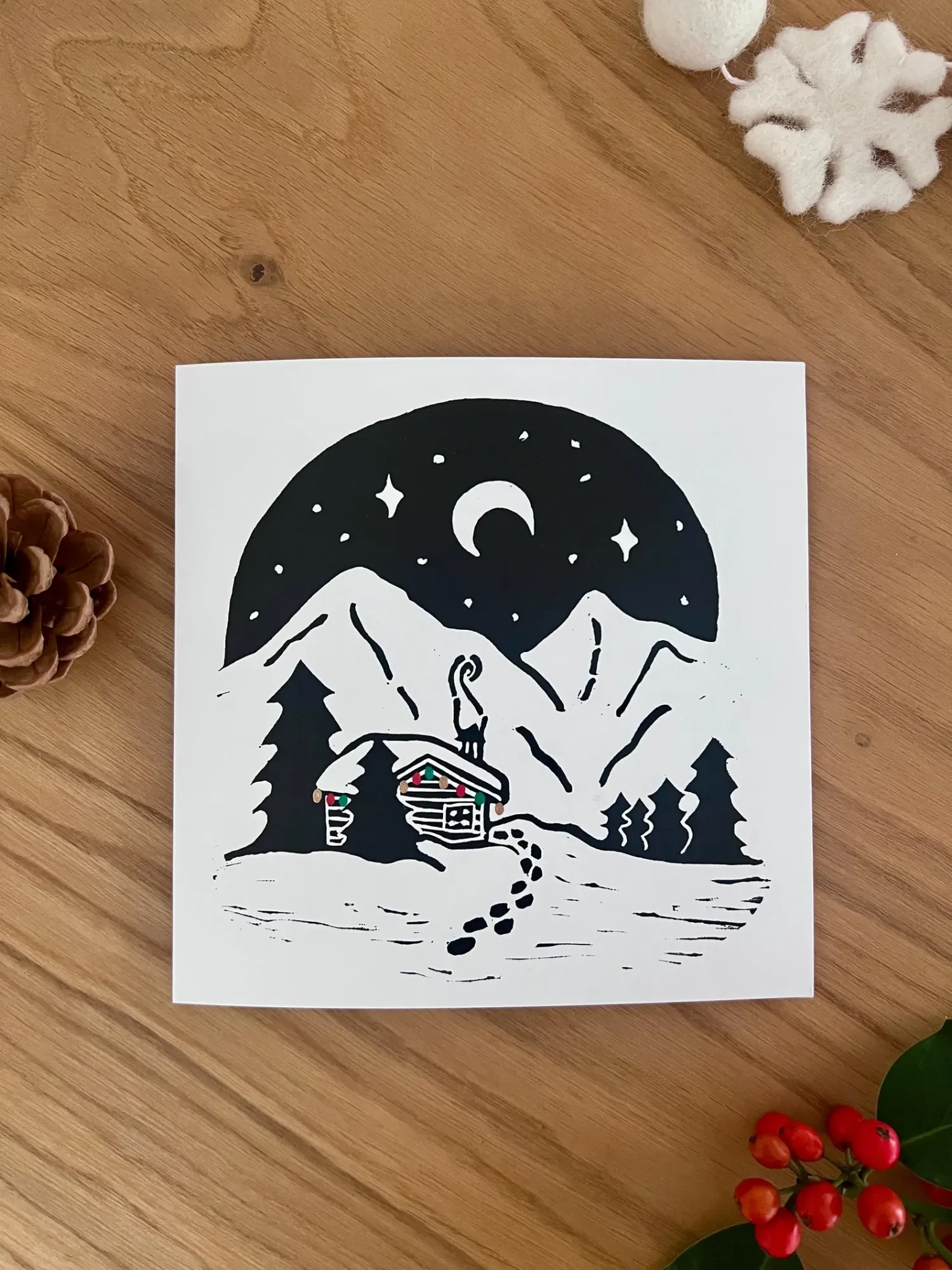 A snowy log cabin with coloured lights and a cat on the chimney, beneath twin mountains