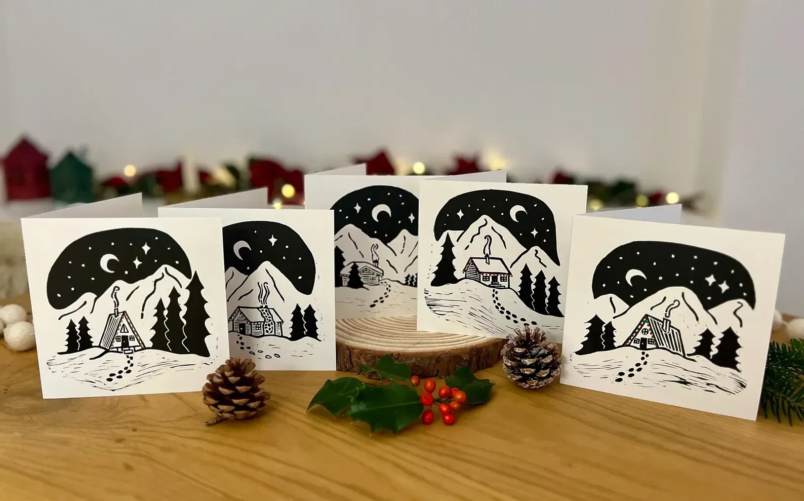 The five hand-printed linocut Christmas cards standing in a row on a wooden table, with pine cones and holly