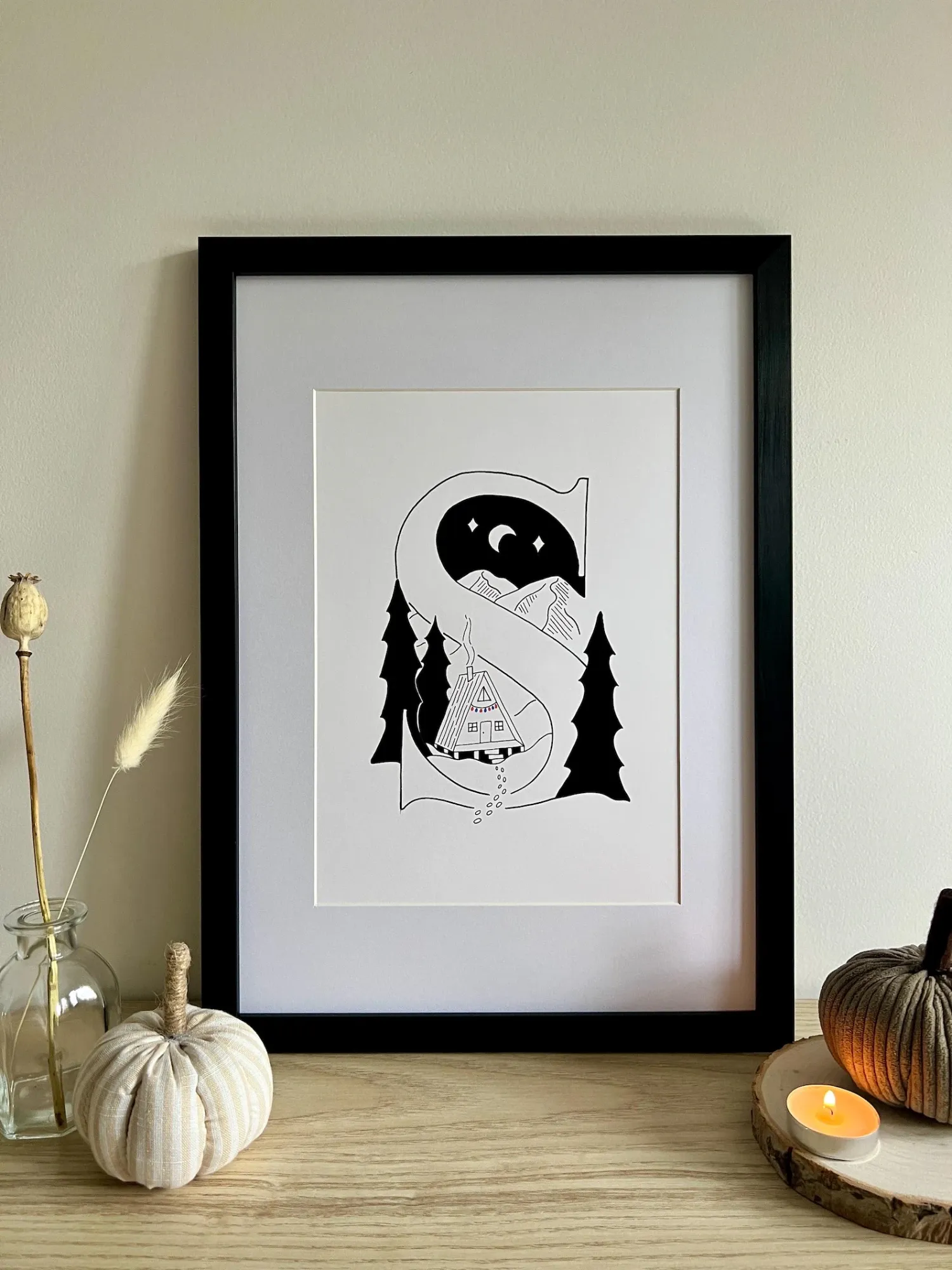 The letter S illustration in a black frame with a white mount, on a shelf beside dried flowers and a candle