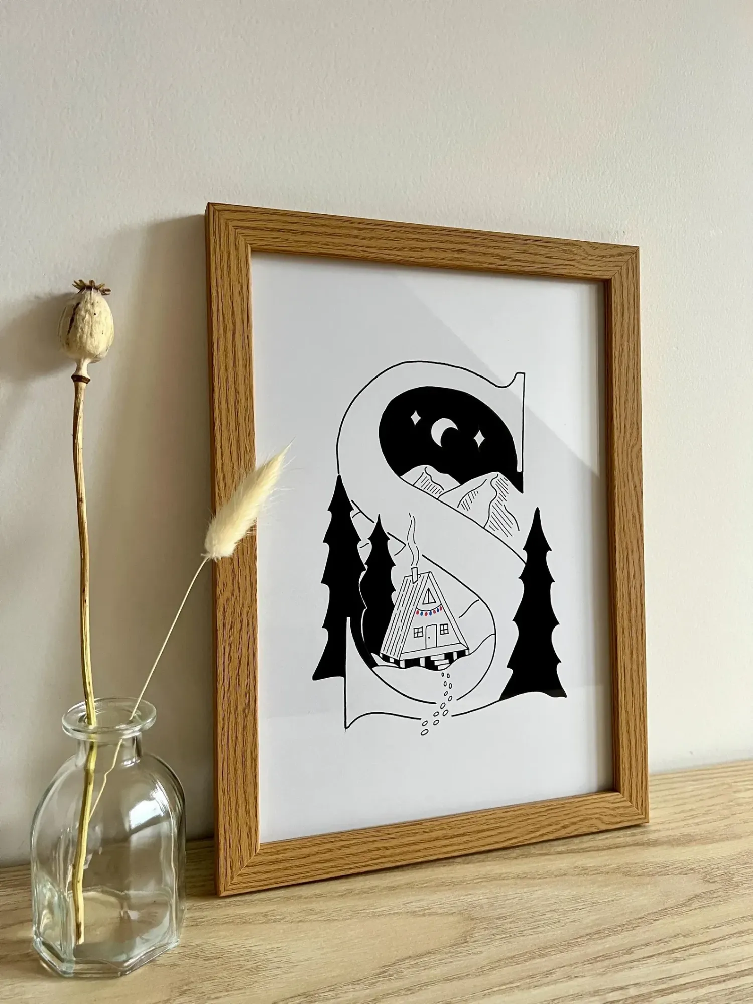 The letter S illustration in an oak frame, leaning against a wall beside a glass bottle of dried grasses
