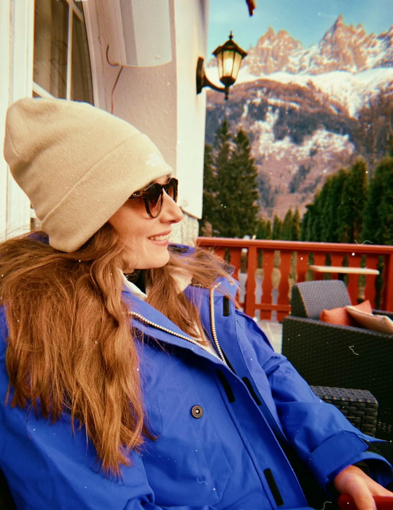 Lydia in a woolly hat and blue coat, smiling on a wooden terrace with snowy mountains behind her